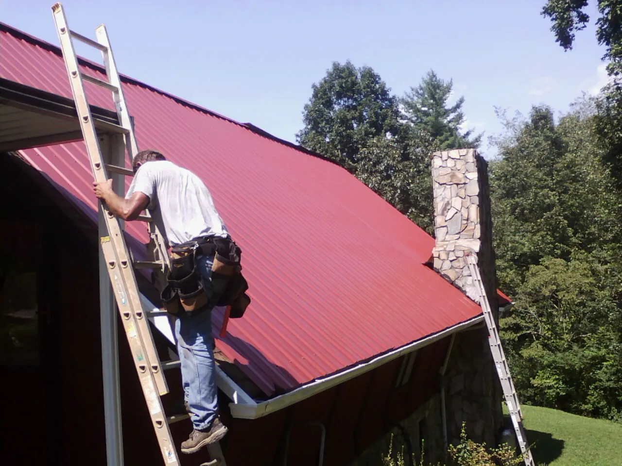 Roofer on a ladder beside a red metal roof and stone chimney — Soffit & Fascia Repair by Roofing Brothers in Hyannis